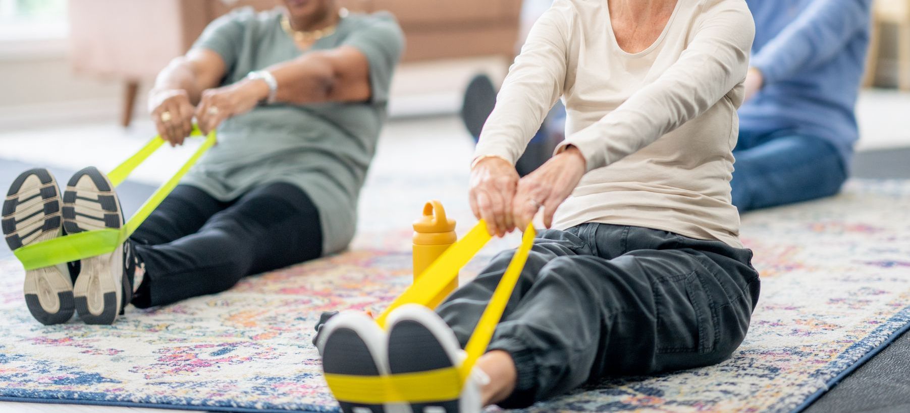 Three people engage in seated exercises using resistance bands on a patterned mat. A water bottle is placed nearby, suggesting a workout session.