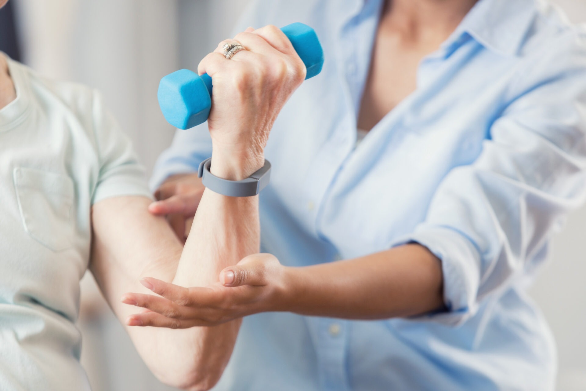 A person assists another in exercising with a blue dumbbell. The focus is on helping with strength training and physical support.