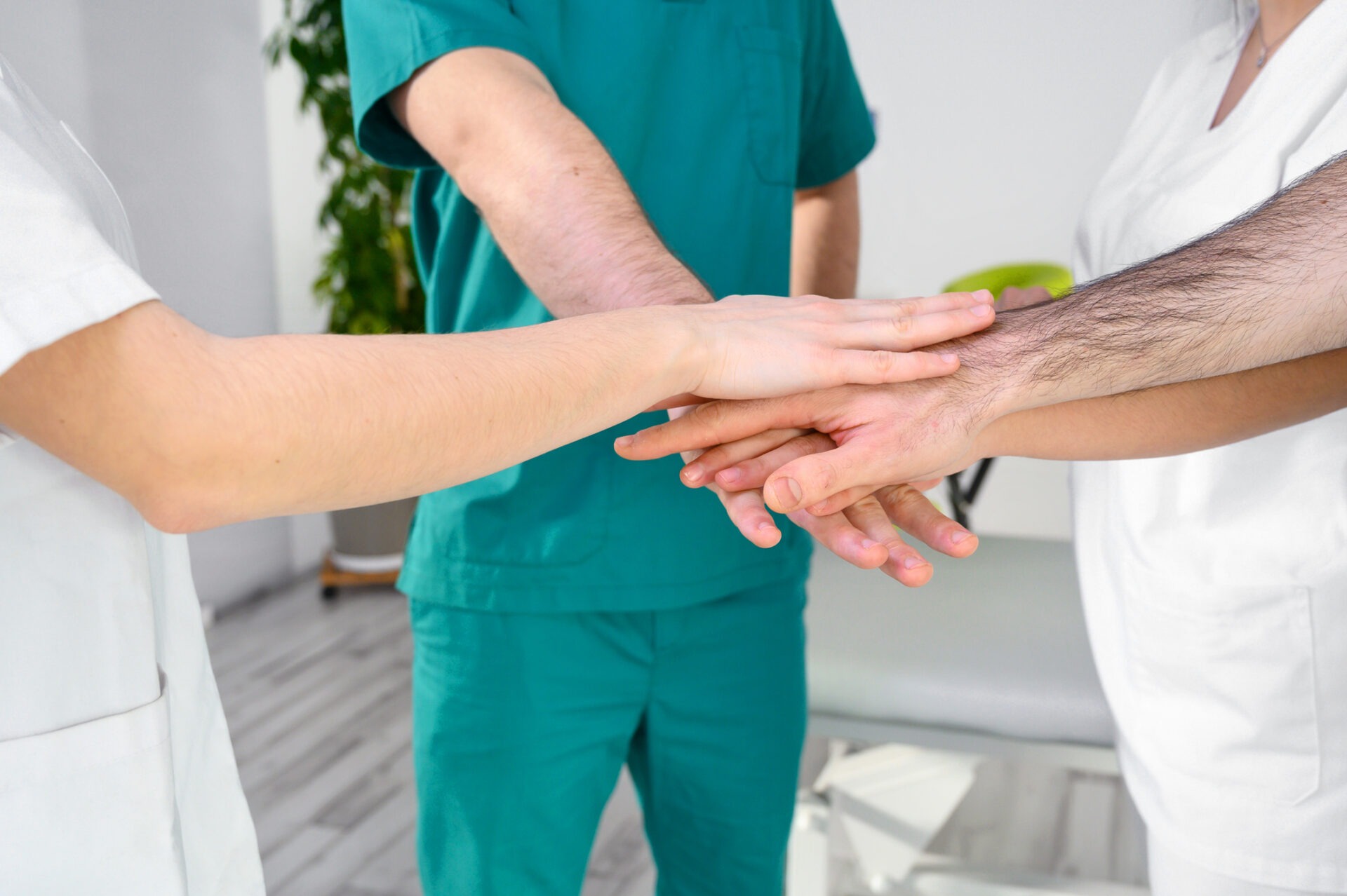 Three people in medical uniforms join hands in a teamwork gesture, standing in a brightly lit room with plants and white walls.