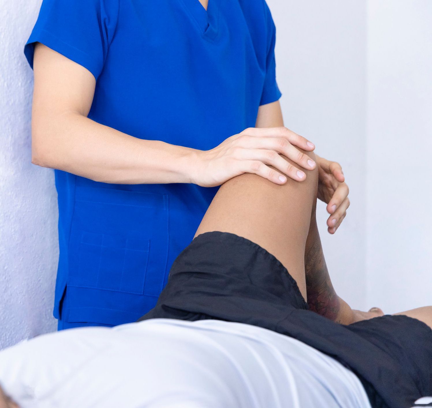 A person in blue scrubs assists another person's leg. The setting appears clinical, indicating a therapeutic or medical scenario, likely physiotherapy or massage.