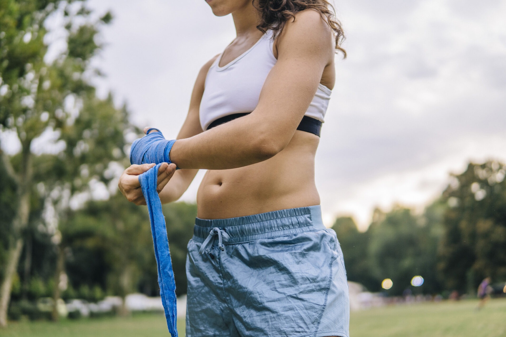 A person outdoors wraps blue sports bandages on their hands, wearing athletic shorts and a top. Green trees and blurred park background are visible.