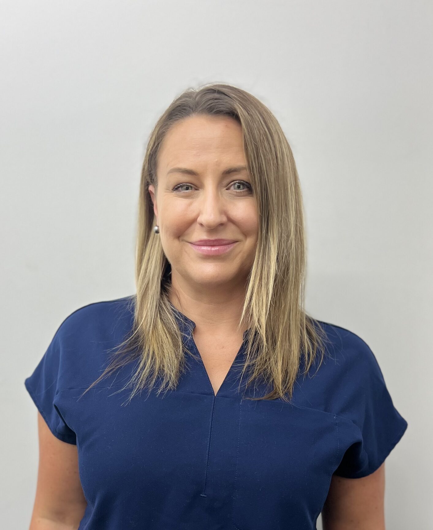 A person with shoulder-length blonde hair is wearing a blue shirt, smiling, standing against a plain white background.