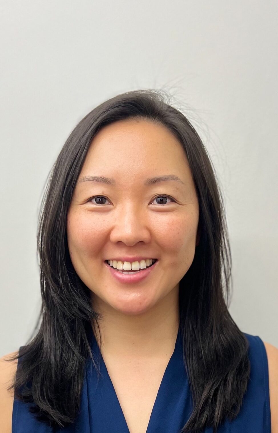 A person with long dark hair smiles against a plain background, wearing a sleeveless blue top, conveying a friendly, approachable demeanor.