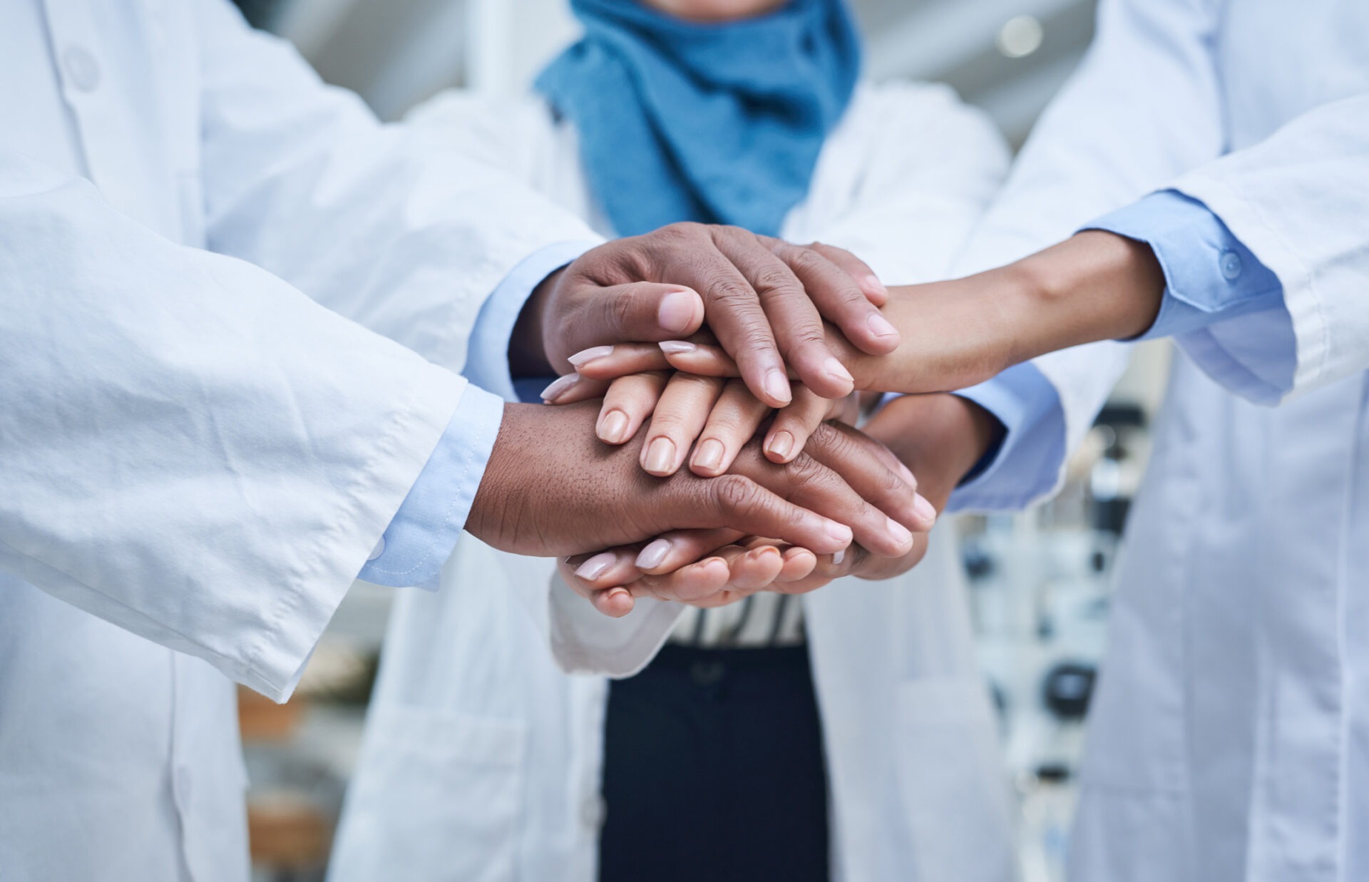 Five people in lab coats unite hands in a supportive gesture. They stand in a bright, modern setting, fostering teamwork and collaboration.