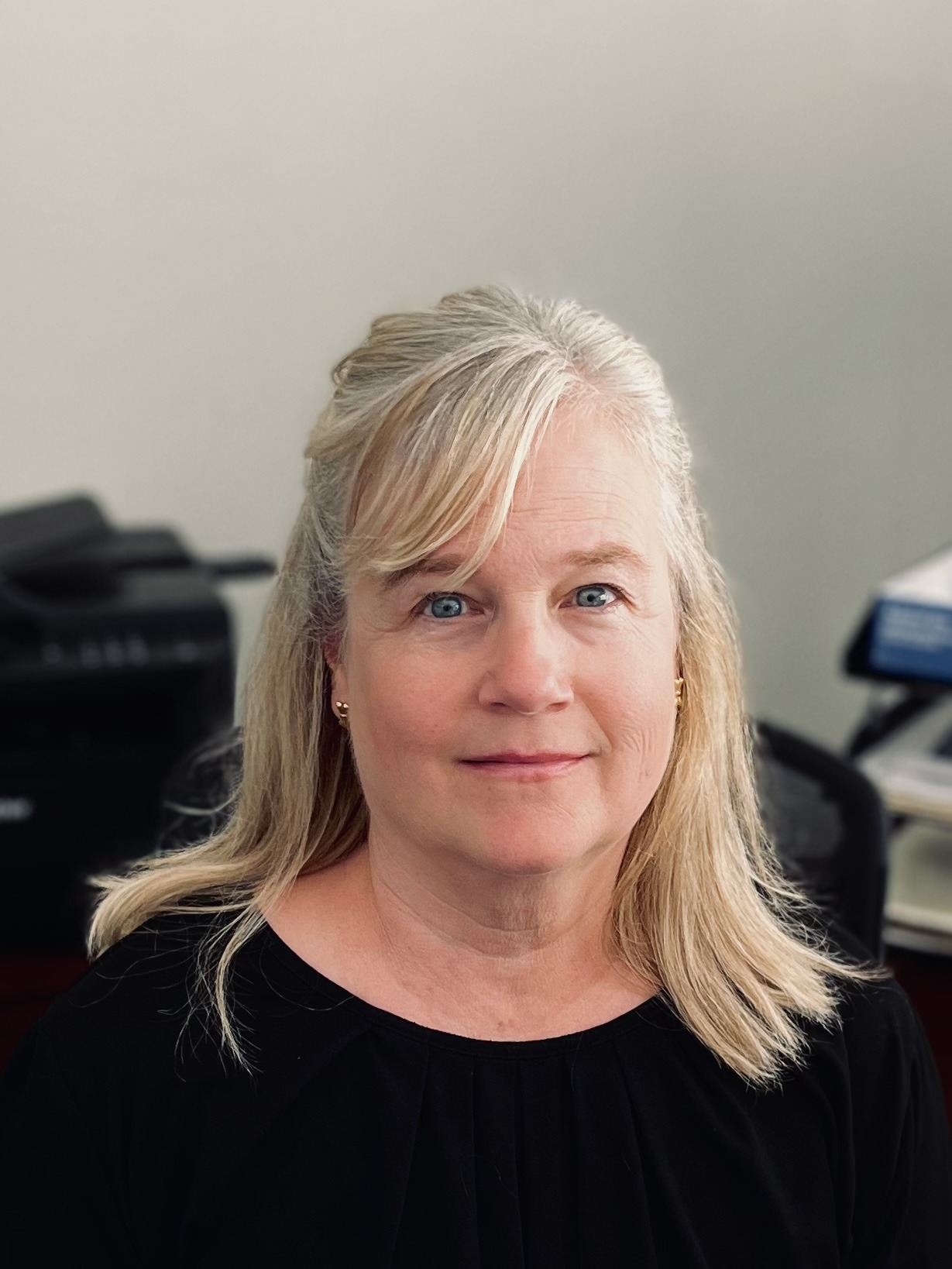 A person with light hair is sitting indoors, wearing a dark top. Background includes a printer and stacked documents.