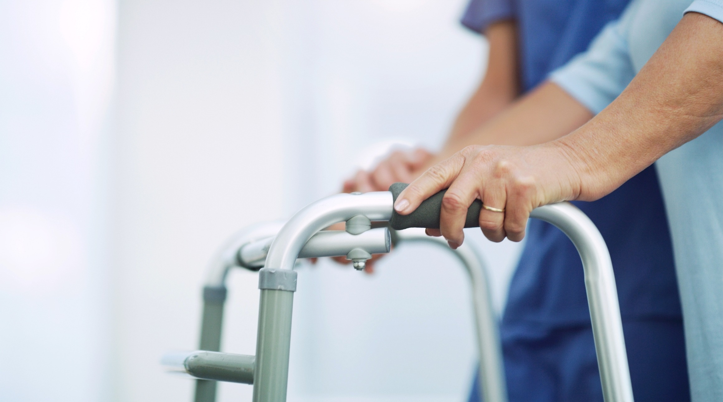 A person uses a walker for support, assisted by another. The focus is on hands and mobility aid, set against a blurred background.