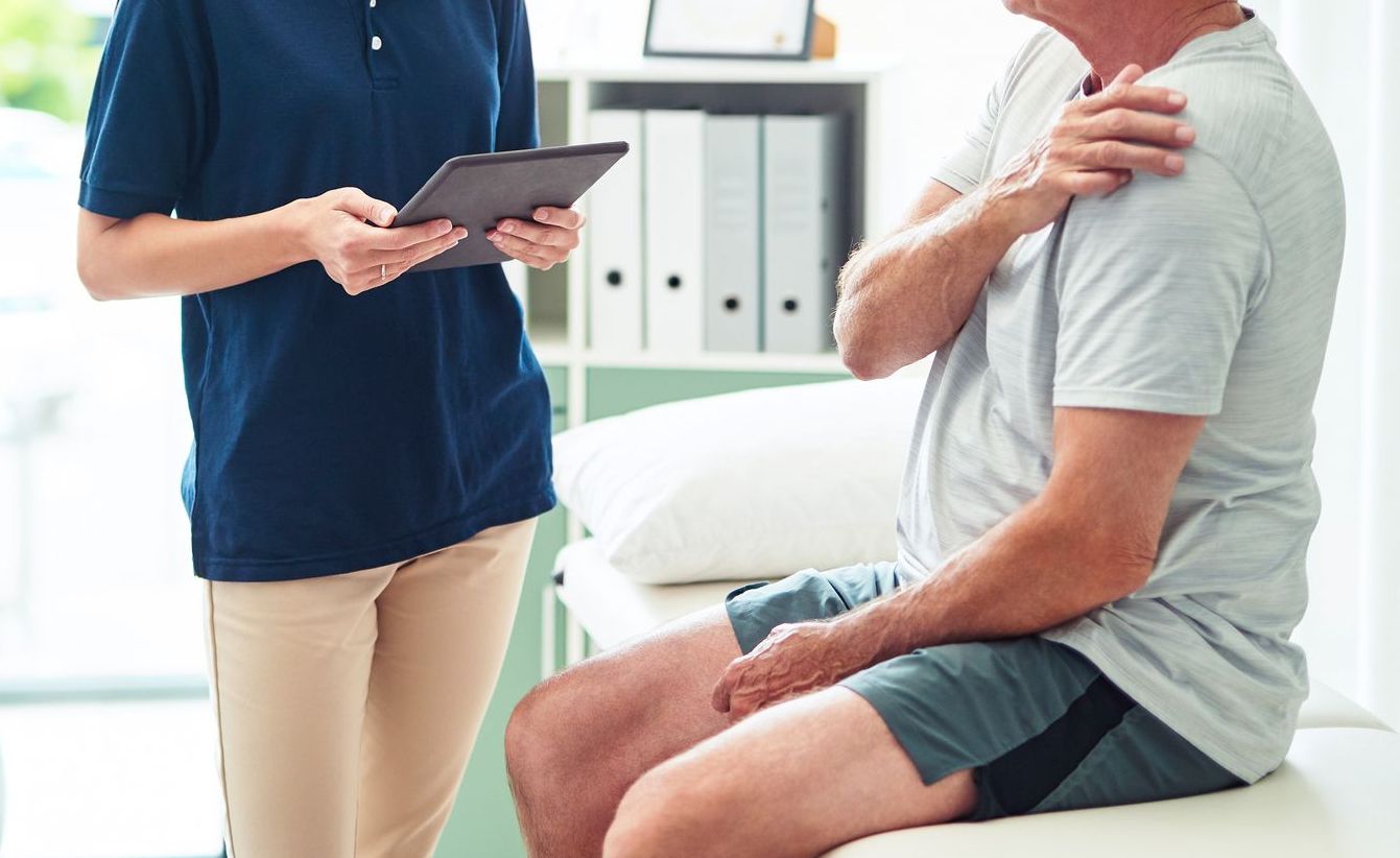 A person sits on a doctor's table, holding their shoulder, while another person stands nearby with a tablet. Medical office setting.