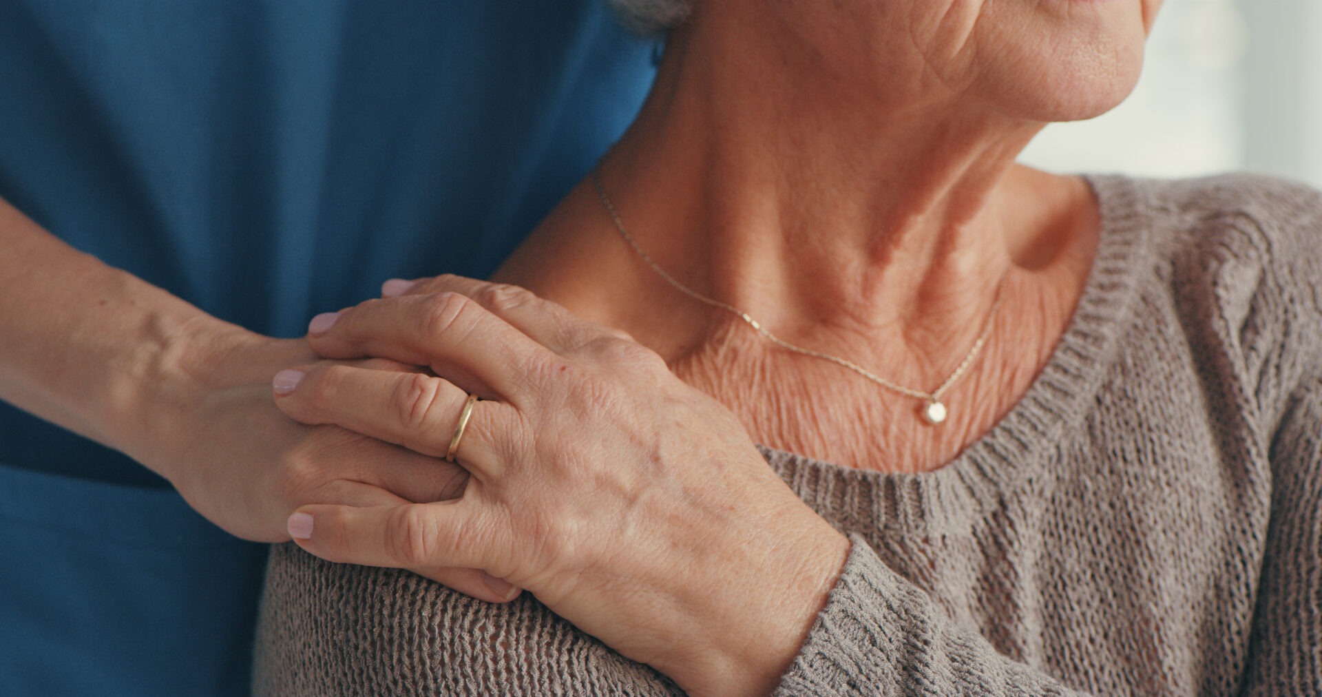 A person gently places a supportive hand on another's shoulder, showing care and empathy. Both individuals wear casual clothing, creating a warm atmosphere.