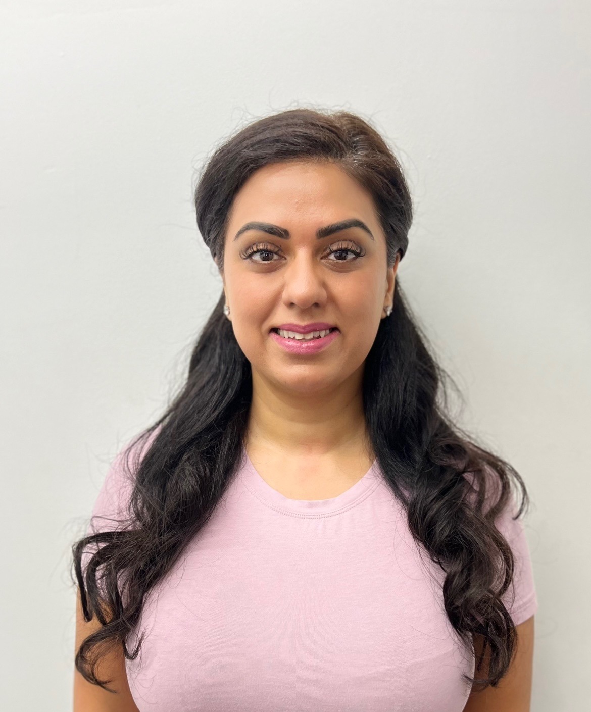 A person with long, dark hair in a pink shirt stands against a plain light background, smiling at the camera.