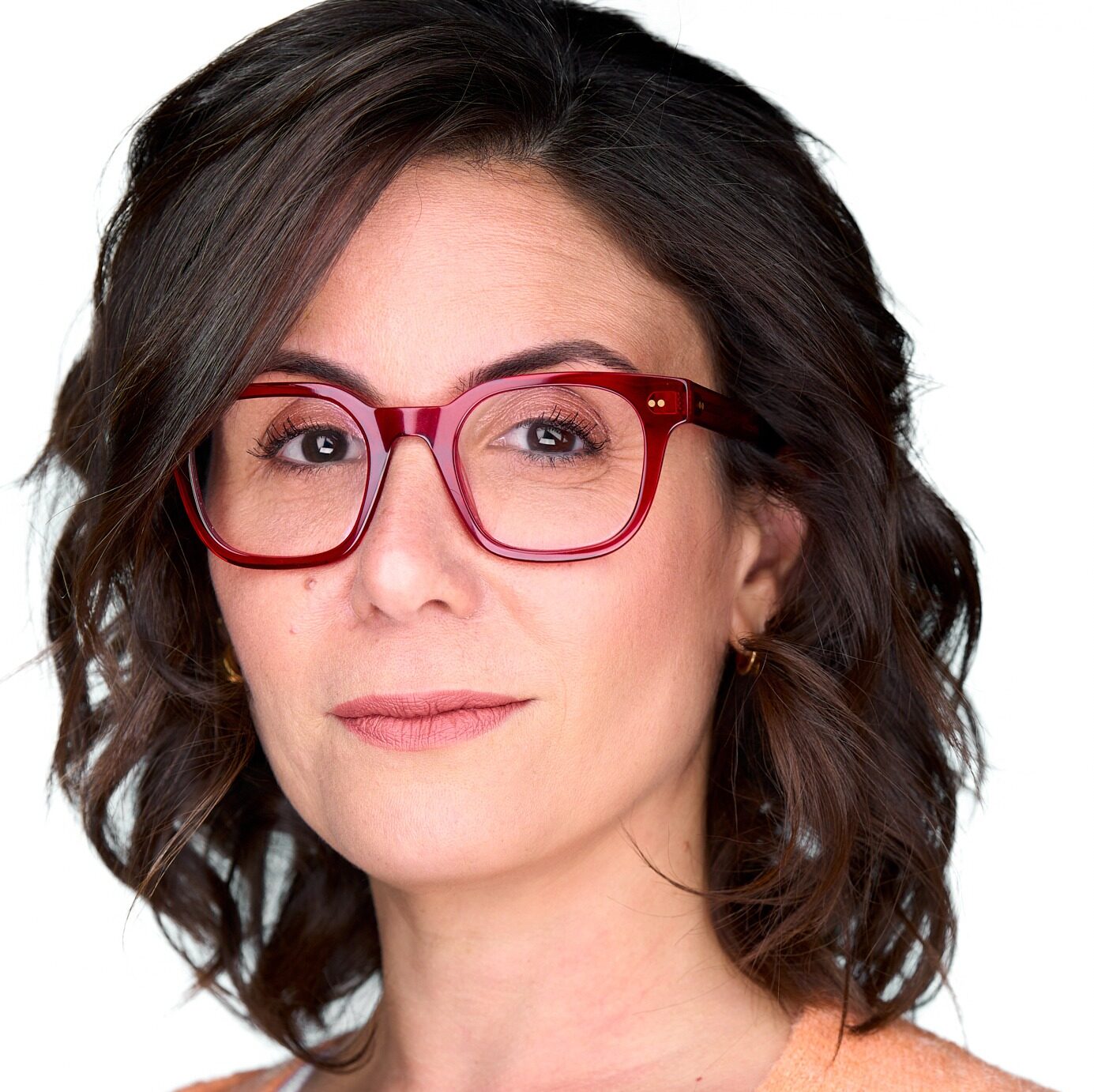 A person with wavy brown hair and red glasses looks directly forward, set against a plain white background.