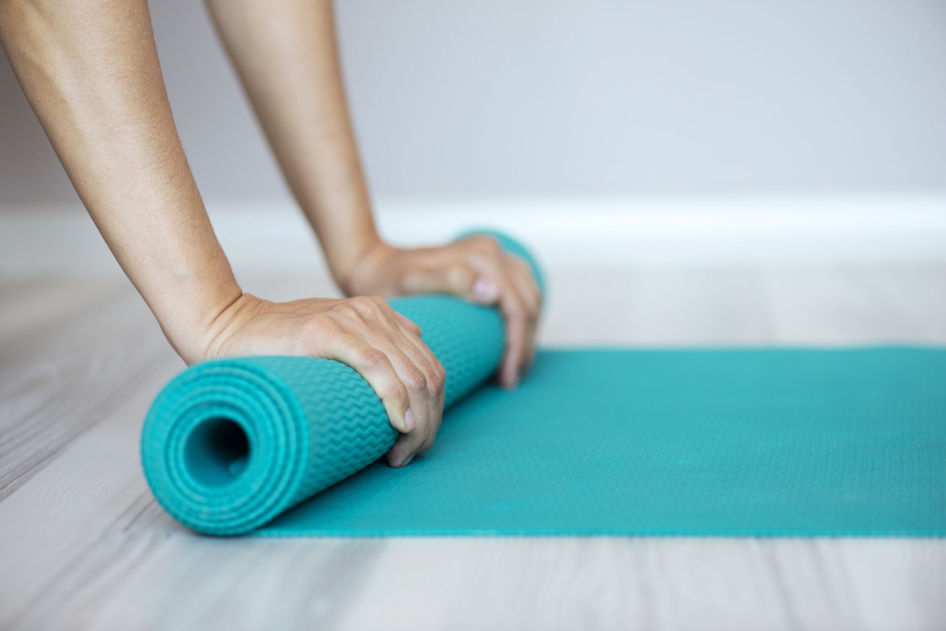 A person rolls up a turquoise yoga mat on a light wooden floor, preparing for exercise or finishing a workout session.