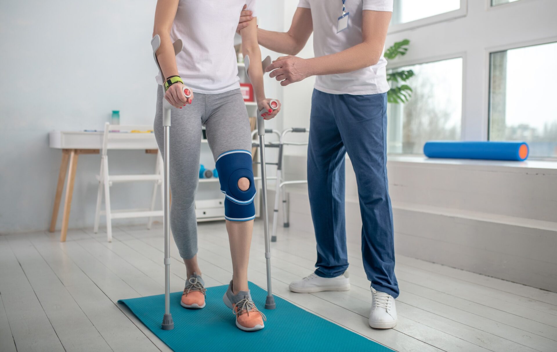 A person with crutches and a knee brace is assisted by another person on a yoga mat in a bright therapy room.