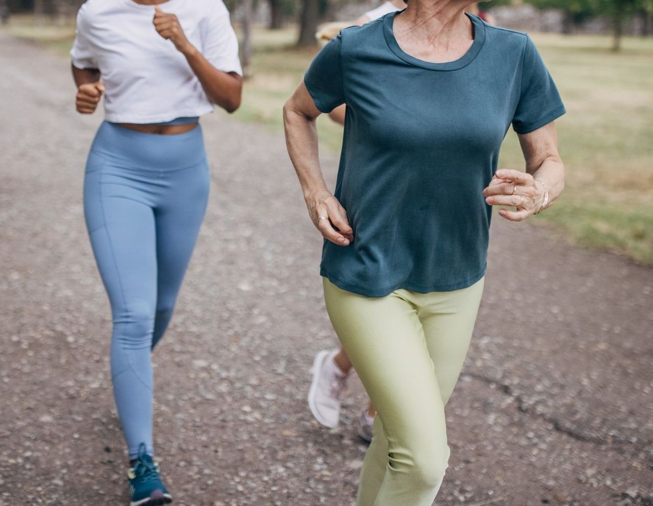 Two people jogging on a gravel path in a park, surrounded by trees. Both are wearing activewear and appear focused on exercise.