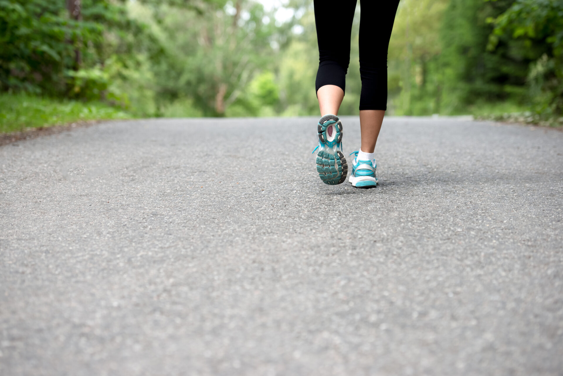 A person wearing athletic shoes jogs on a tree-lined path, with lush greenery on both sides and no visible landmarks or buildings.