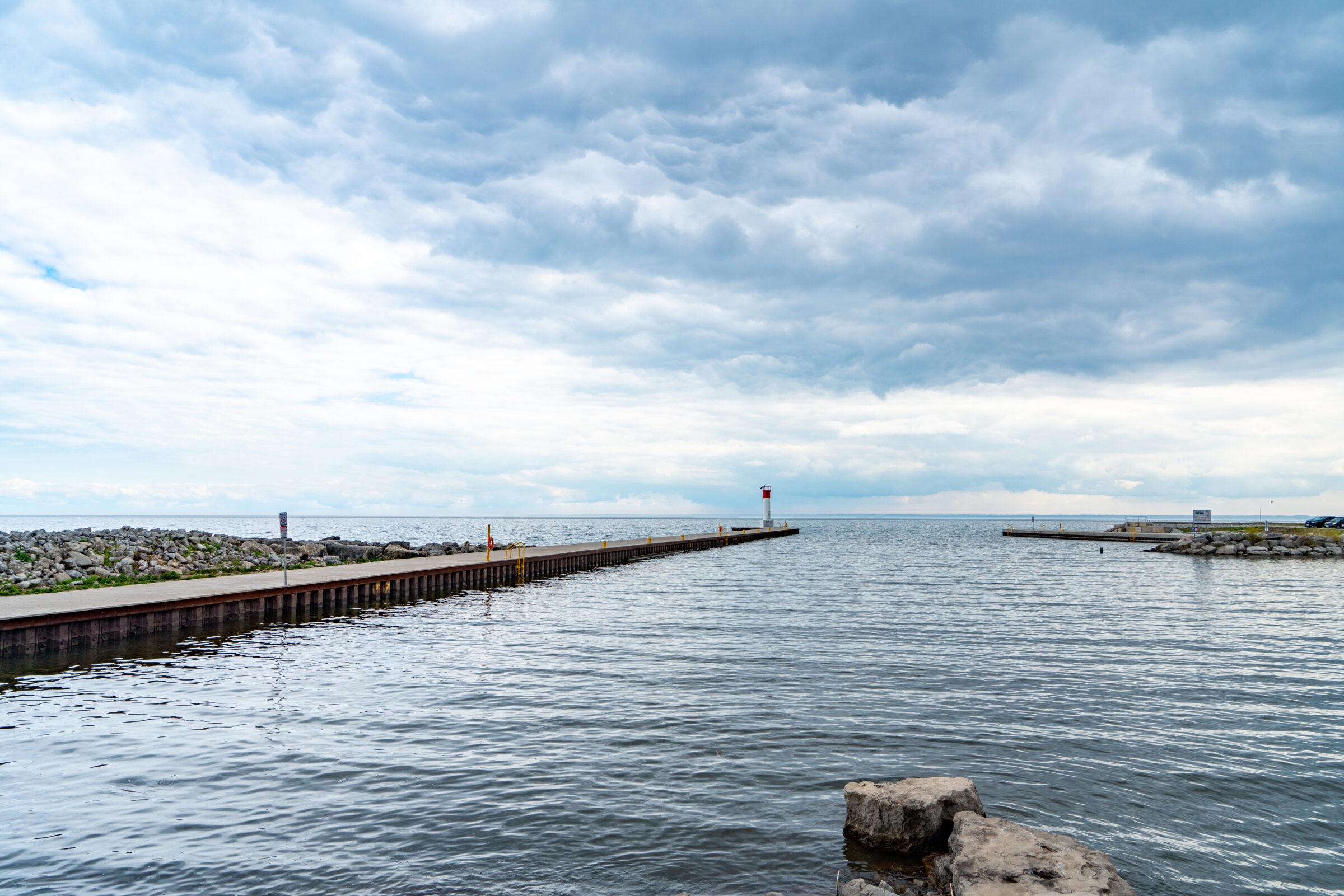 A calm waterfront scene with a rocky shoreline and cloudy sky; pier with red marker extends into the water. No landmarks visible.