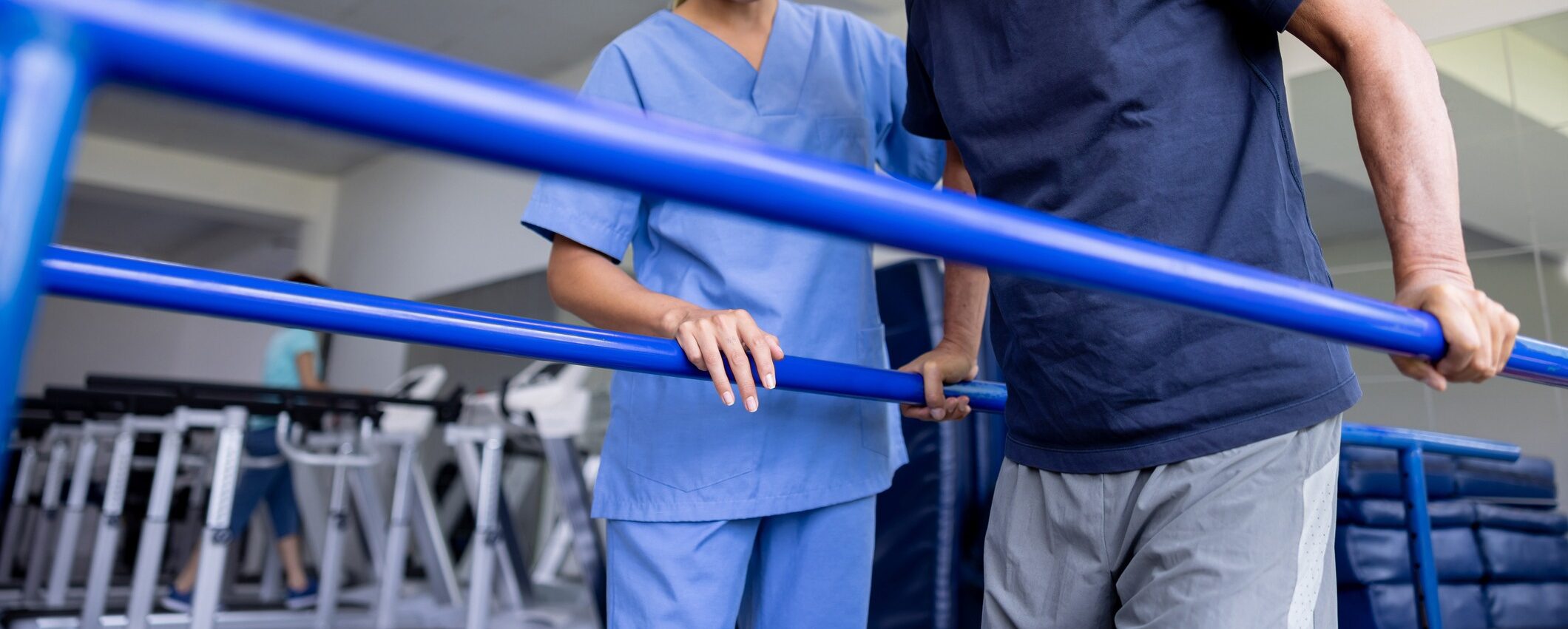 A person in blue scrubs assists another person during rehabilitation exercises, using parallel bars in a clinical setting with exercise equipment nearby.