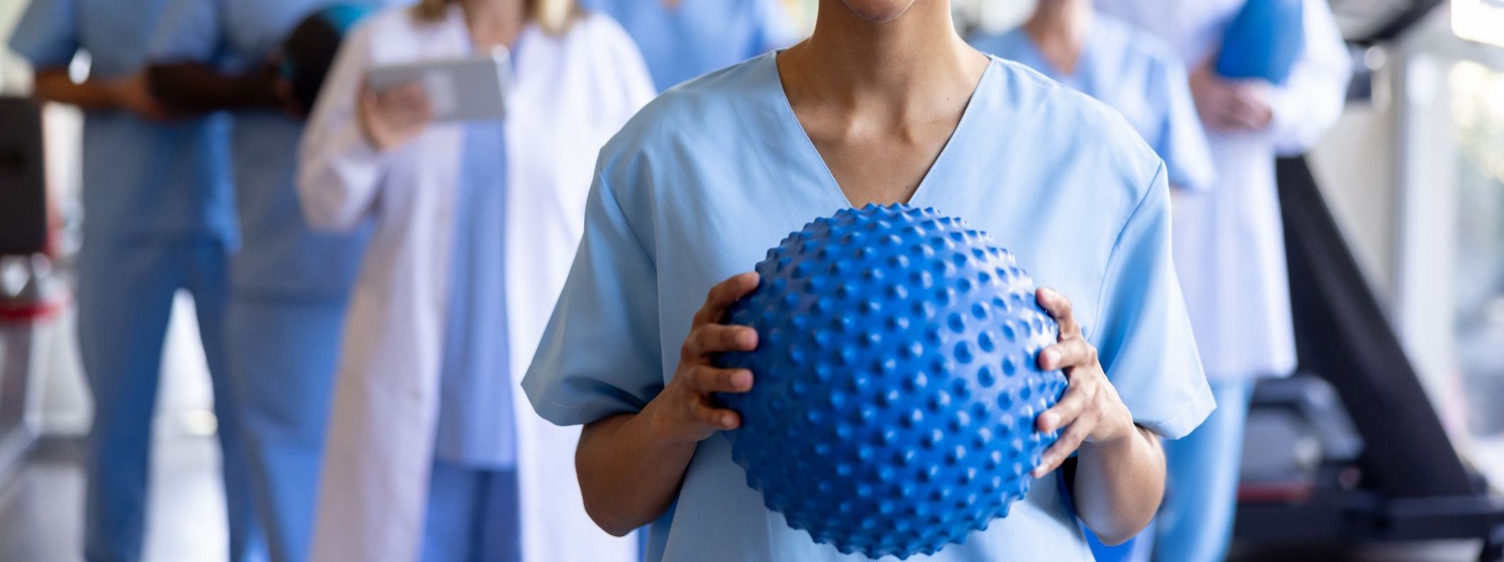 A person in scrubs holds a blue spiky exercise ball, with other medical professionals in the background, suggesting a healthcare setting.