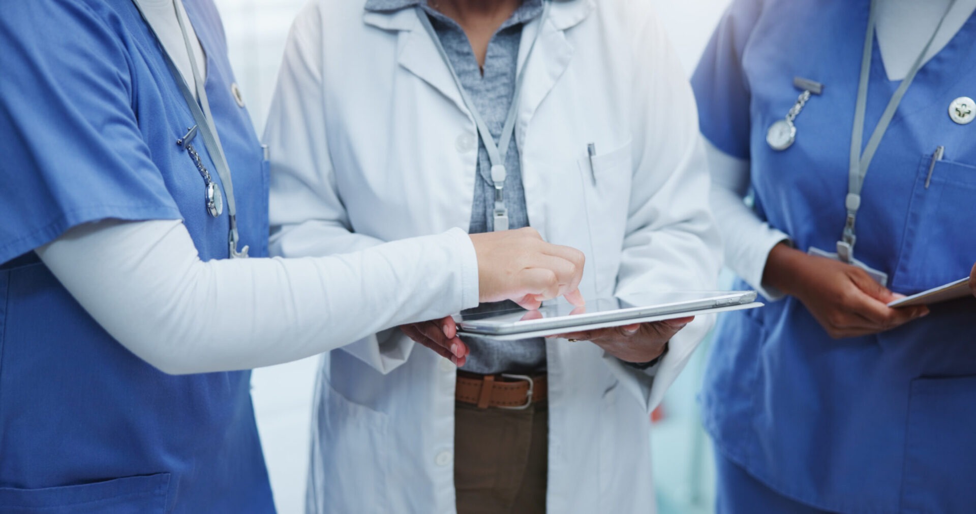 Three healthcare professionals in scrubs and lab coats discuss patient information on a tablet in a hospital setting, focusing on collaborative care.