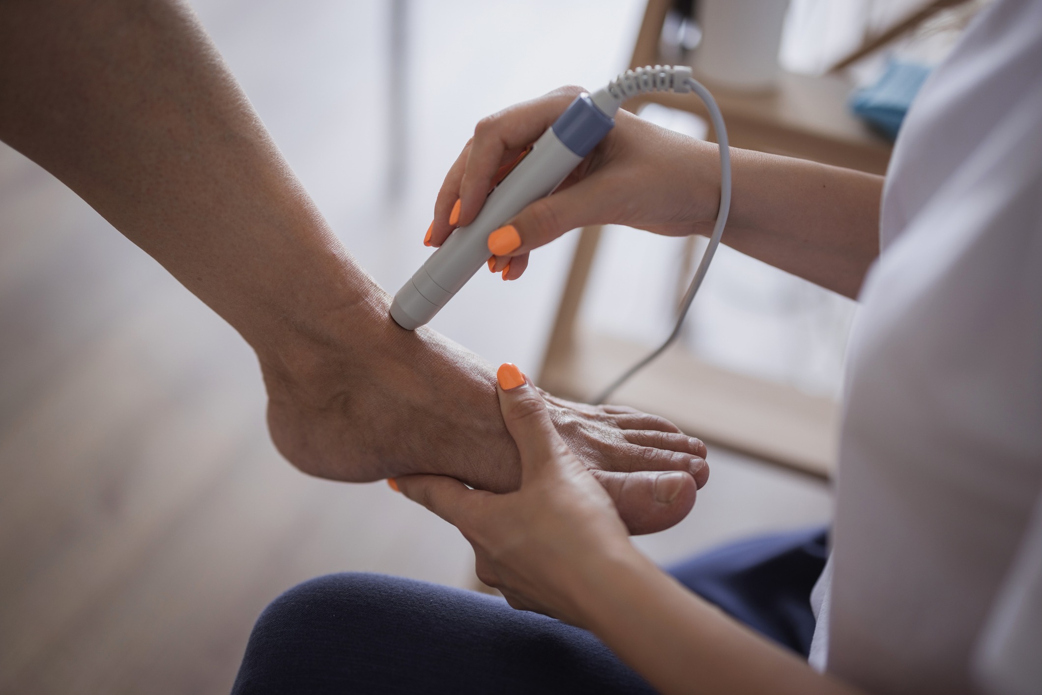 A person uses a handheld device on another person's foot, possibly for a medical or therapeutic procedure, in a softly lit room.