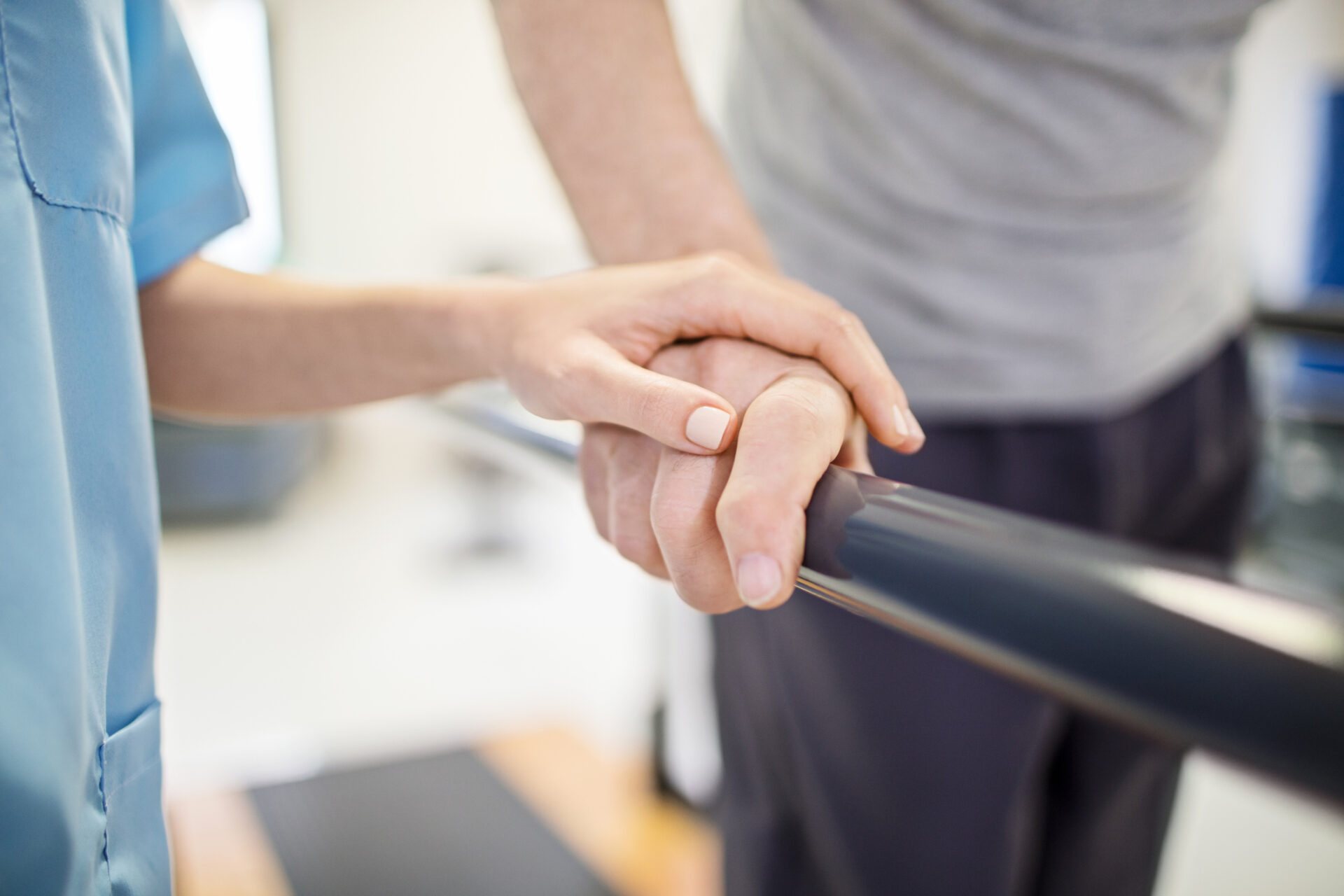 A person assists another with physical therapy, holding their hand on a parallel bar in a rehabilitation setting. Close-up view emphasizes support.