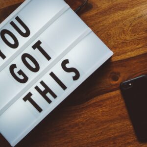 A lightbox with "YOU GOT THIS" sits on a wooden table beside a smartphone and a partly visible laptop with a floral case.