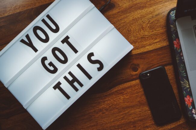 A lightbox with "YOU GOT THIS" sits on a wooden table beside a smartphone and a partly visible laptop with a floral case.