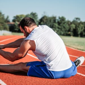A person on a track, stretching with one leg extended. The background features trees and a clear sky.