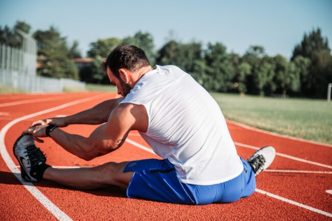 A person on a track, stretching with one leg extended. The background features trees and a clear sky.