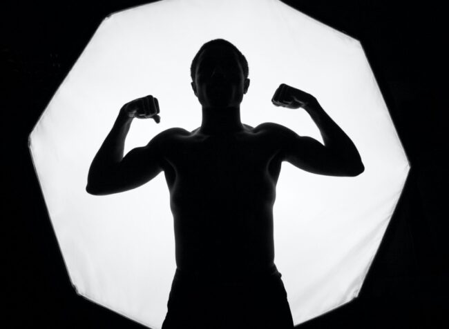 Silhouetted person flexing muscles in front of bright octagonal light source, creating a dramatic, high-contrast image in black and white.